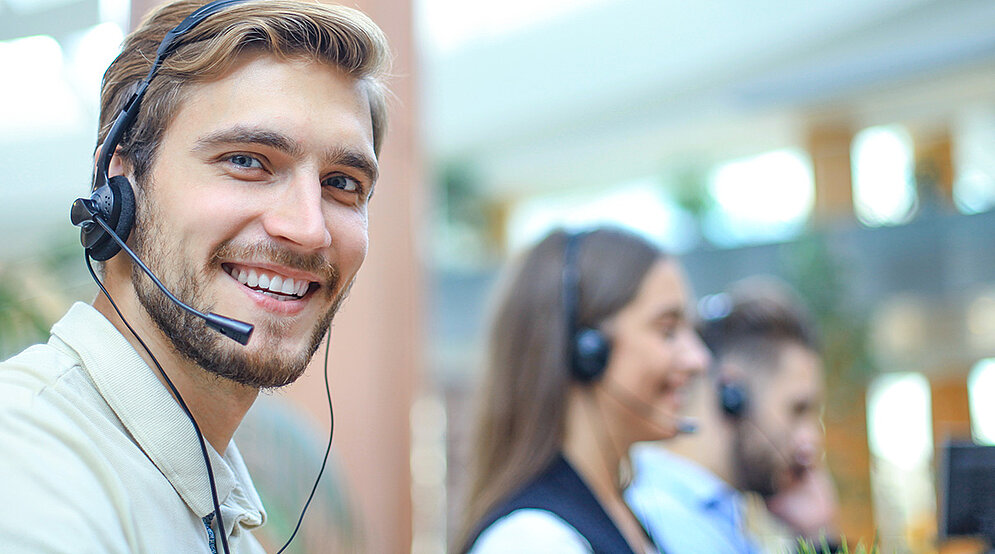 Ein männlicher Service-Mitarbeiter Mitte 30 trägt ein Headset und schaut freundlich lachend in die Kamera. Im Hintergrund ist eine weibliche Service-Kollegin zu sehen, die schmunzelnd in ihr Headset spricht.