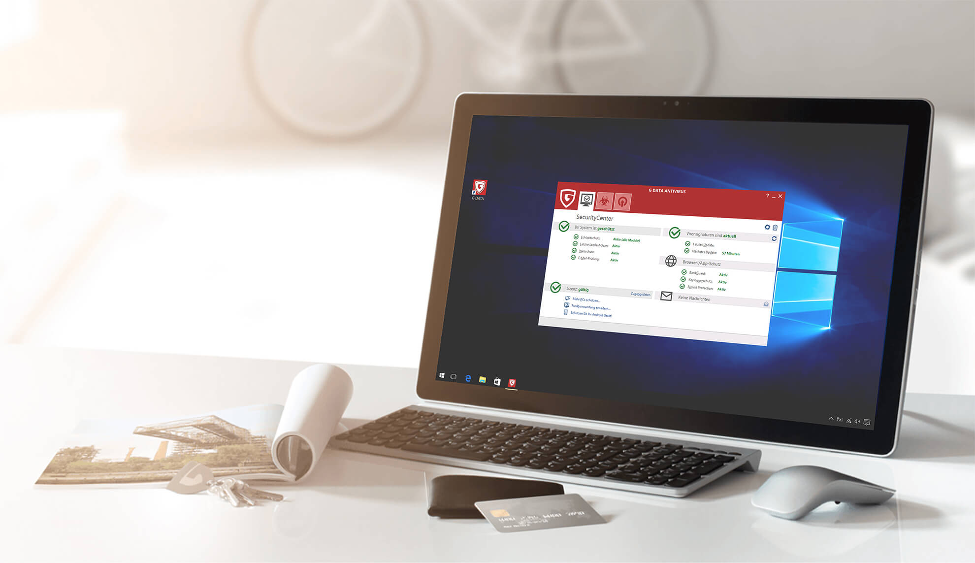 Auf dem Foto erkennt man einen Schreibtisch. Auf diesem befinden sich ein Magazin, ein Schlüssel, ein Portmonnaie mit Kreditkarte und ein Bildschirm mit Tastatur und Maus. Auf dem Display ist die Softwareoberfläche der G DATA Antivirus für Windows zu sehen. 
